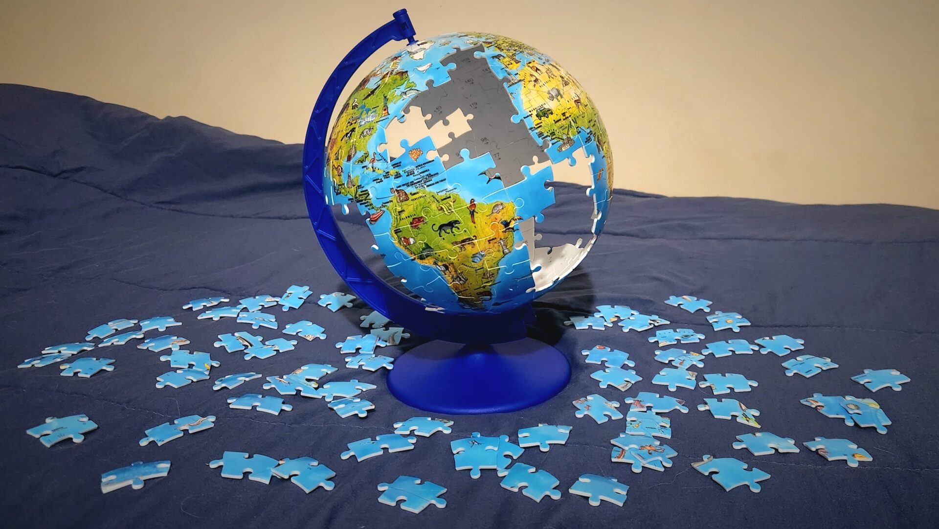 A mostly assembled puzzle globe precariously mounted on the globe stand with huge gaps in the puzzle where oceans should be, with the remaining pieces scattered around the stand on a bed.