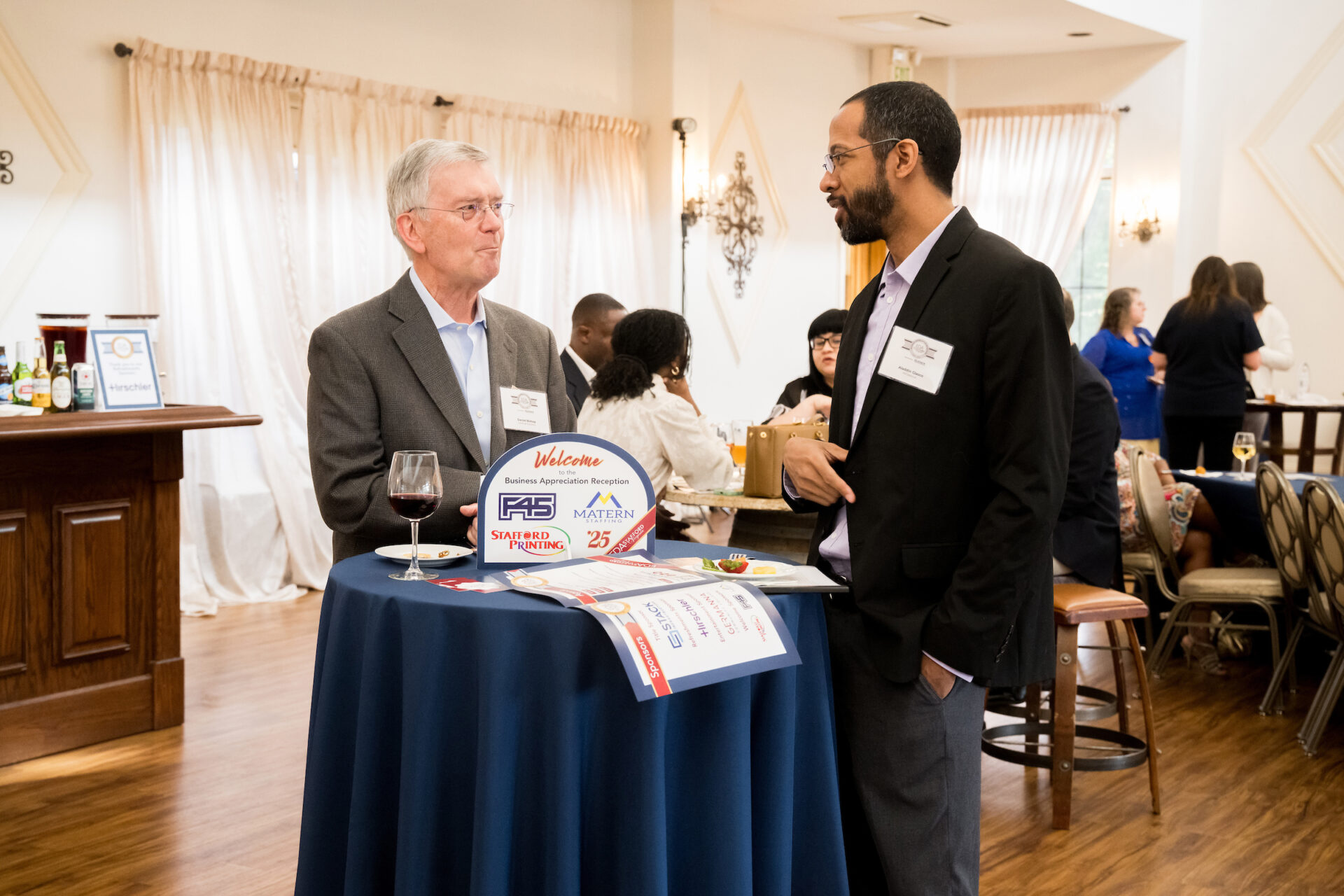 Daniel Bishop (left), CEO of DanEuB Solutions, stands in conversation with Aladdin Glasco (right), founder of MrGlasco.ai, at a small round table during the 2025 Stafford County Business Appreciation Reception at Potomac Point Winery. The table is covered with event flyers and a welcome sign displaying sponsor logos. Other attendees are seated and mingling in the warmly lit background.