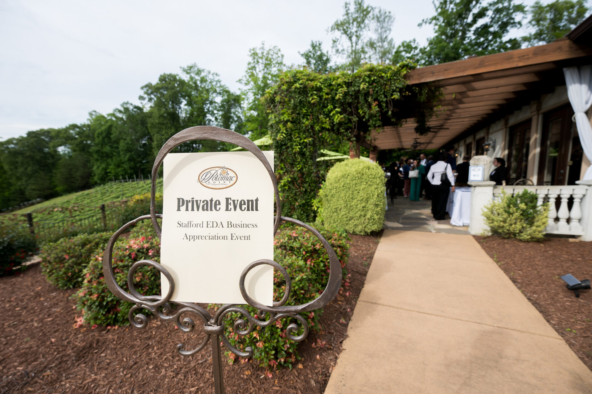 A sign reading “Private Event – Stafford EDA Business Appreciation Event” stands at the entrance to Potomac Point Winery, framed by manicured landscaping and flower beds. In the background, attendees are gathered under a vine-covered pergola near the entrance, where event staff welcome guests at a check-in table.
