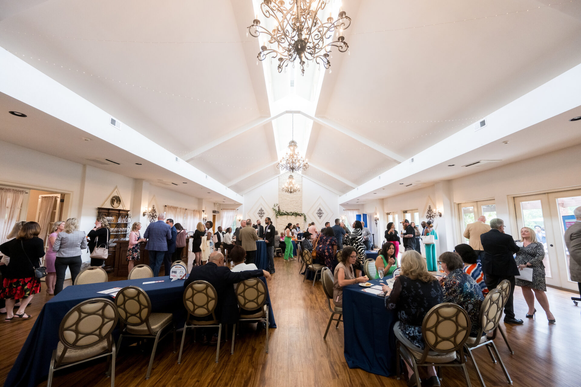 Wide-angle view of the 2025 Stafford County Business Appreciation Reception, showing a warmly lit event hall filled with business professionals networking, conversing, and seated at tables. Elegant chandeliers hang from the high vaulted ceiling, while natural light streams through tall windows along the right side of the room. Attendees in formal and business casual attire engage in lively discussions throughout the venue.